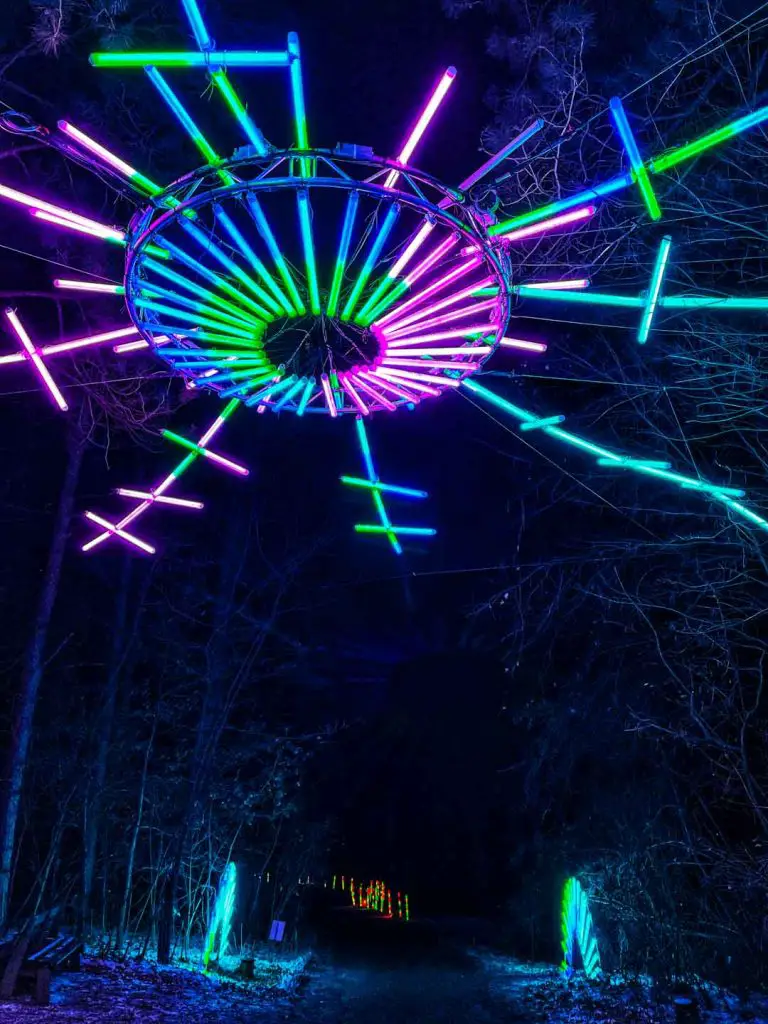 A neon snowflake light display hangs above the trail in the illuminated forest of Glenlore Trails in Commerce Township, Michigan, USA