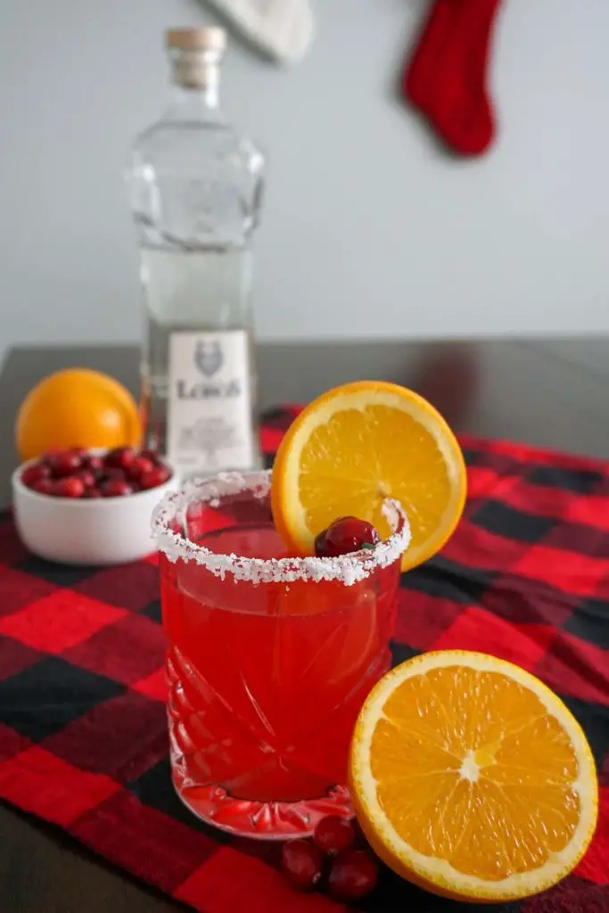 Cranberry Orange Margarita holiday cocktail in a salt-rimmed old fashioned cocktail glass and garnished with an orange slice and fresh cranberries pictured in a Christmasy holiday setting with stockings hanging in background, red-and-black buffalo check cloth on brown wooden table, and Lobos 1707 Tequila Joven bottle and cocktail garnishes