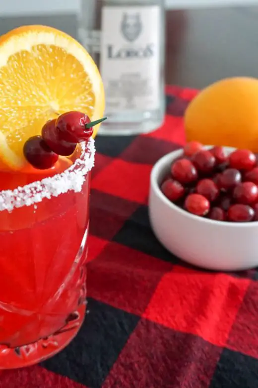 Cranberry orange margarita holiday cocktail in a salt-rimmed old fashioned cocktail glass in a Christmas holiday setting with red-and-black buffalo check cloth, Lobos 1707 Tequila Joven bottle, halved navel orange, and small bowl of fresh cranberries for cocktail garnish