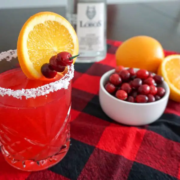 Cranberry orange margarita holiday cocktail in a salt-rimmed old fashioned cocktail glass in a Christmas holiday setting with red-and-black buffalo check cloth, Lobos 1707 Tequila Joven bottle, halved navel orange, and small bowl of fresh cranberries for cocktail garnish