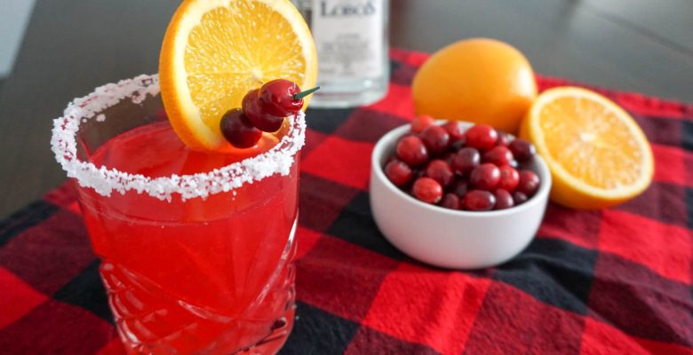 Cranberry orange margarita holiday cocktail in a salt-rimmed old fashioned cocktail glass in a Christmas holiday setting with red-and-black buffalo check cloth, Lobos 1707 Tequila Joven bottle, halved navel orange, and small bowl of fresh cranberries for cocktail garnish
