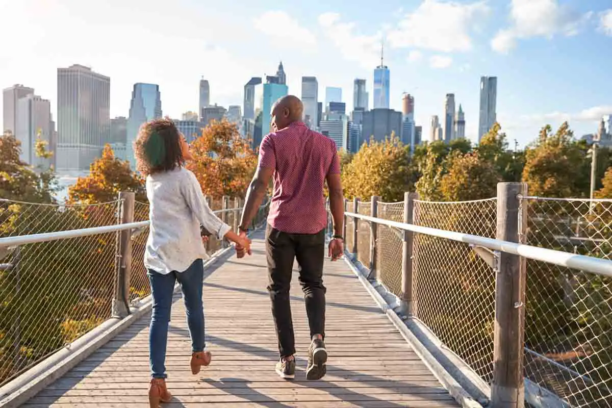 Couple walks hand-in-hand along path with New York City skyline in the background