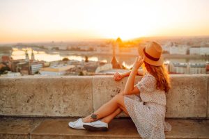 Female traveler wearing sneakers, dress, and hat admires a European city view at sunrise