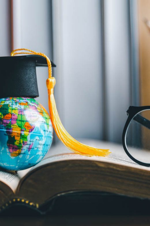 Study abroad concept with graduation cap (mortar board and tassel) on mini globe on a student's desk with open book, reading glasses, and container of writing utensils in background