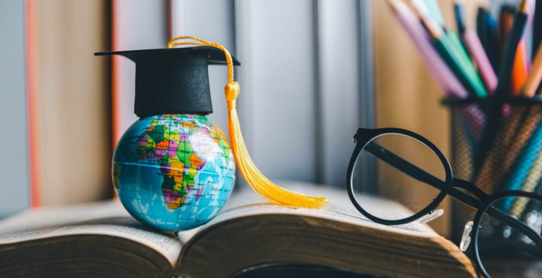 Study abroad concept with graduation cap (mortar board and tassel) on mini globe on a student's desk with open book, reading glasses, and container of writing utensils in background