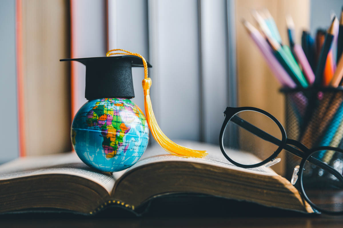 Study abroad concept with graduation cap (mortar board and tassel) on mini globe on a student's desk with open book, reading glasses, and container of writing utensils in background