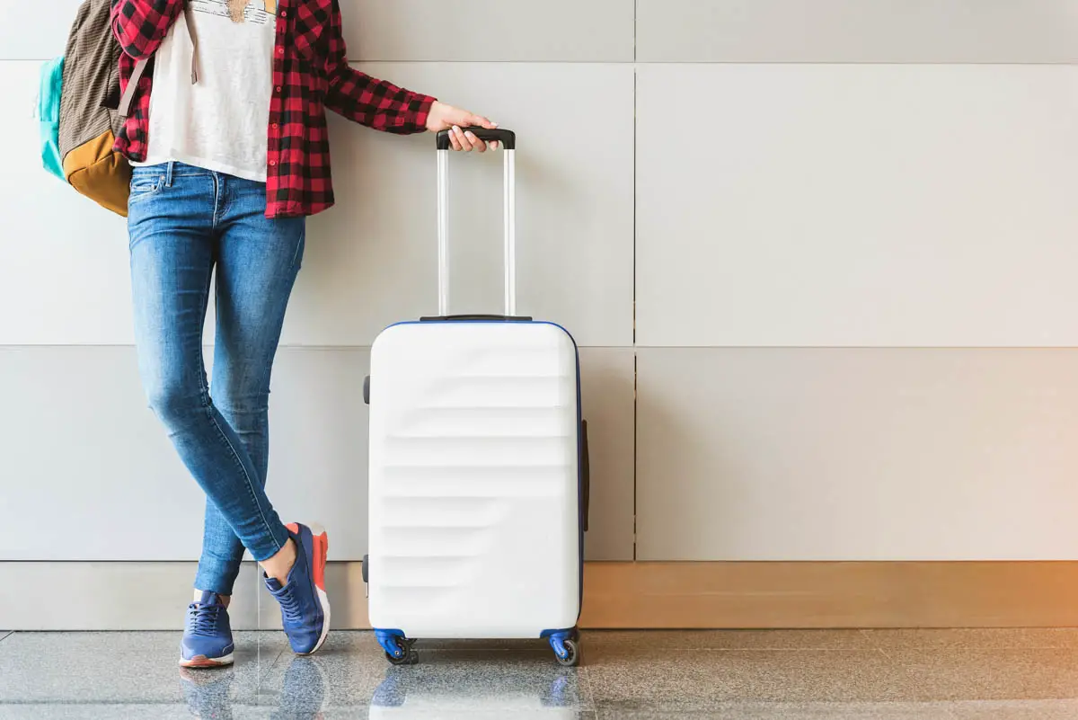 Young female traveler dressed in casual clothing poses at airport with hard-side spinner suitcase and backpack