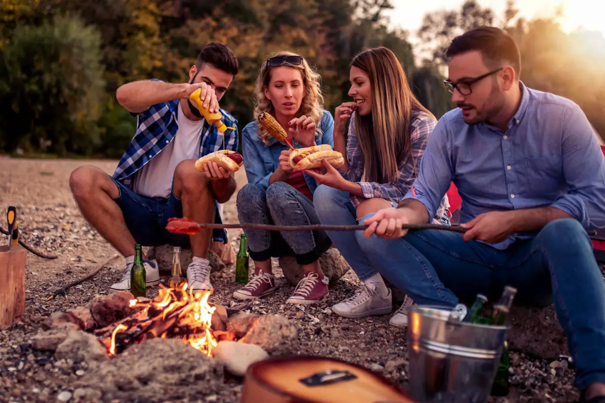 Group of young adults roasting and eating easy camping foods, including hot dogs, around a campfire (© Ivanko80/AdobeStock)