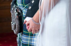 A bride and groom, clad in a tartan kilt and sporran, hold hands at a Scottish wedding