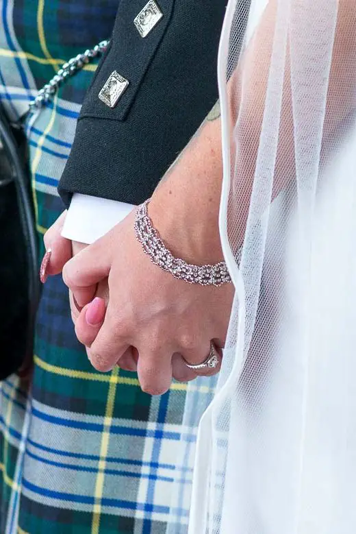 A bride and groom, clad in a tartan kilt and sporran, hold hands at a Scottish wedding