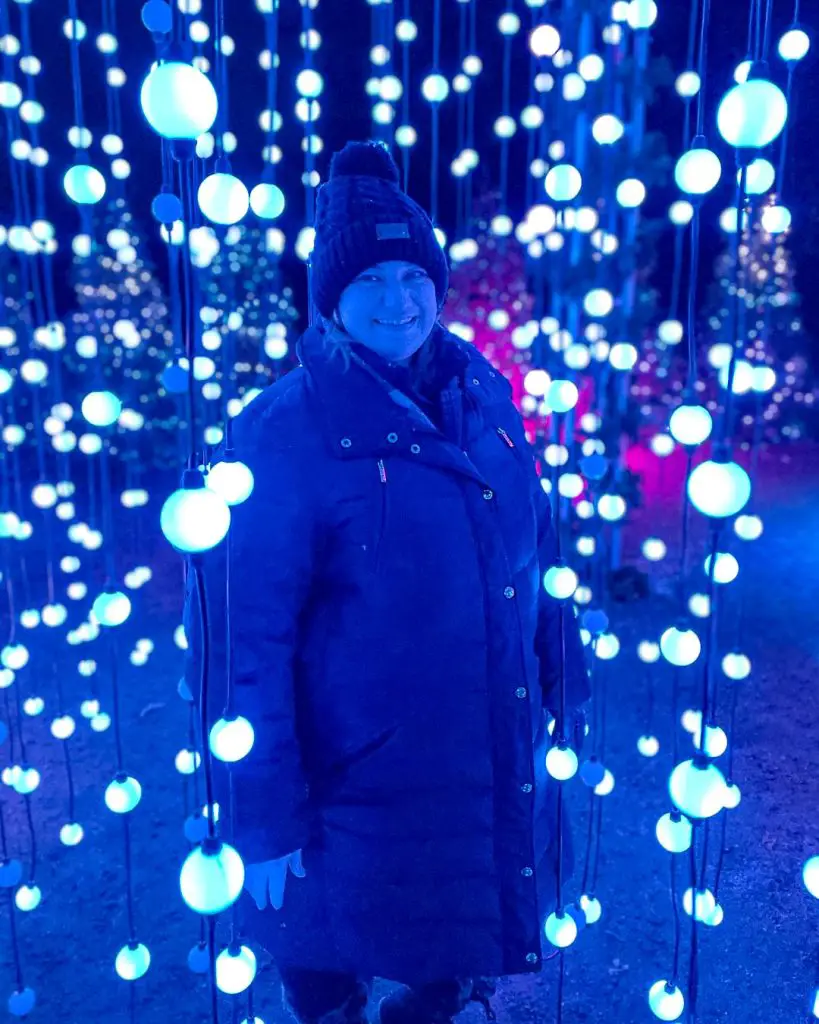 Woman stands amid hanging lights at Glenlore Trails Aurora, an illuminated forest holiday lights trail walk, in Commerce Township, Michigan, USA