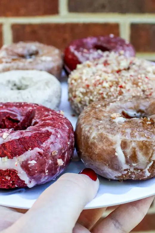 A woman's hand holds a white plate with half a dozen cake doughnuts, including red velvet donuts, Dutch crumb, powedered sugar, and glazed donuts
