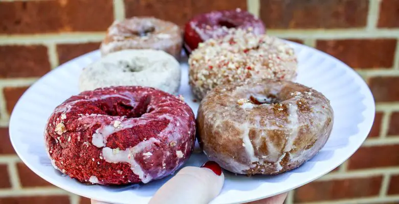 A woman's hand holds a white plate with half a dozen cake doughnuts, including red velvet donuts, Dutch crumb, powedered sugar, and glazed donuts