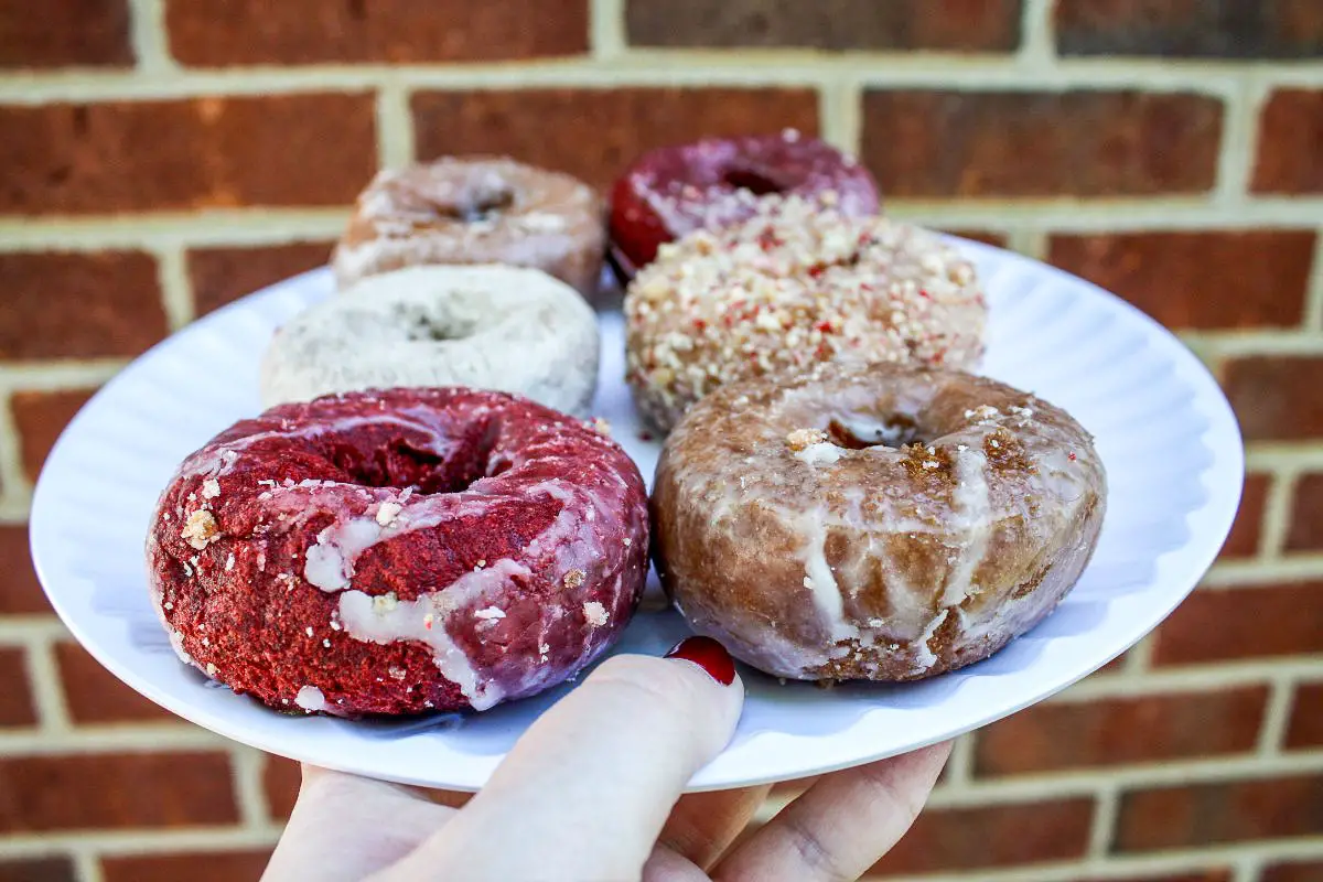 A woman's hand holds a white plate with half a dozen cake doughnuts, including red velvet donuts, Dutch crumb, powedered sugar, and glazed donuts