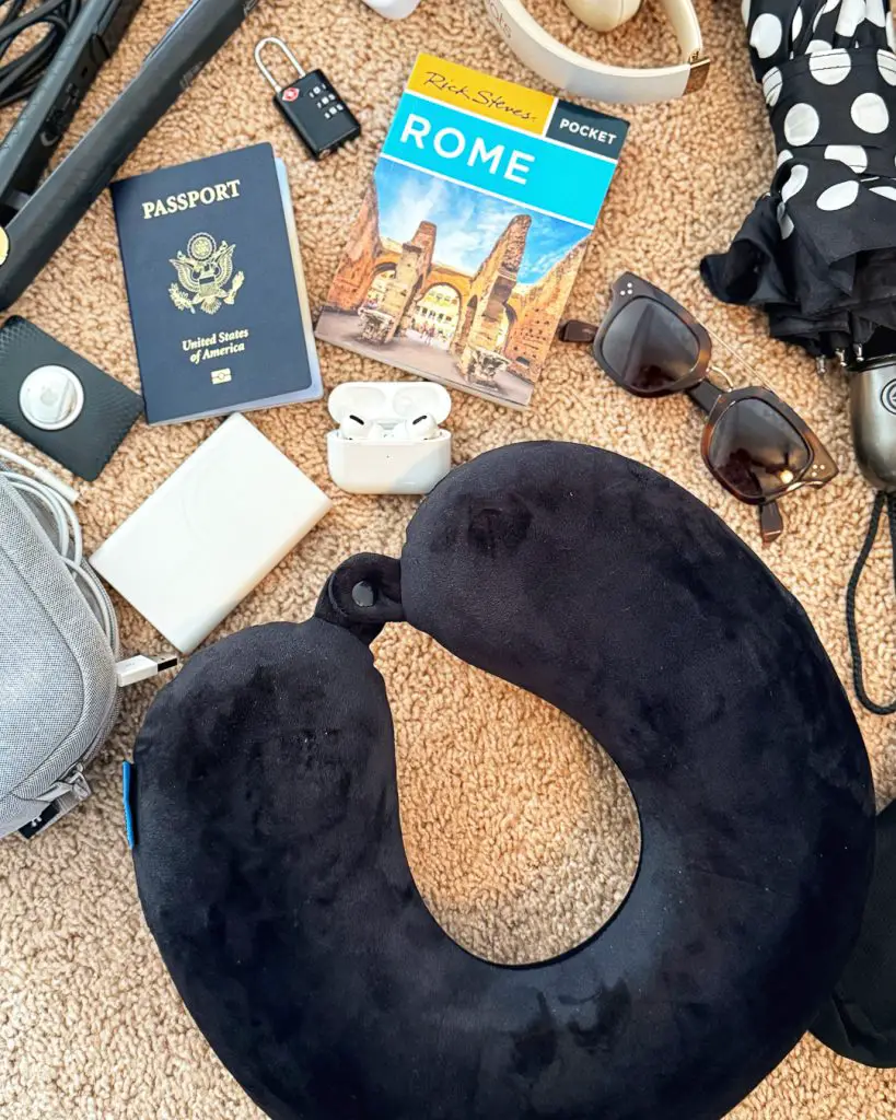 Europe travel essentials including a Brookstone travel neck pillow, AirTag, power bank, Airpods, sunglasses, umbrella, tech organizer, passport, and pocket guidebook